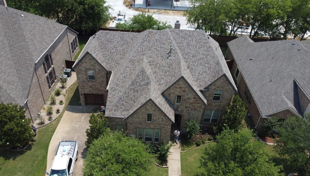 Aerial view of a large brick house with a complex shingle roof, driveway, and front yard landscaping.