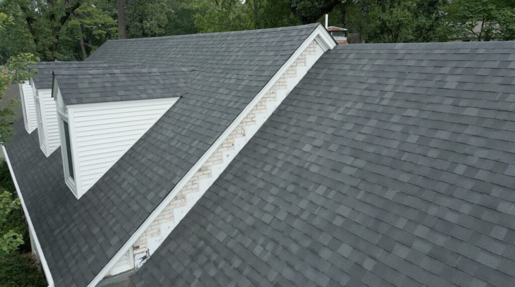 Close-up view of a residential roof with dark gray asphalt shingles and white dormer windows surrounded by trees.